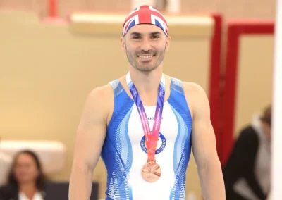 Coach Stefan Kolimechkov smiling with a medal after a gymnastics championship in the United Kingdom.