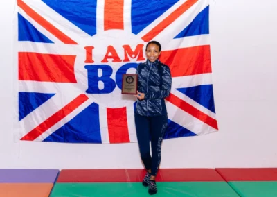 Club's Deputy welfare officer Mary-Anne holding an award plaque in front of a British Gymnastics flag at Chelsea Gymnastics Academy Christmas event in Kensington.