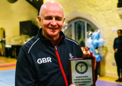 Club Welfare Officer smiling and holding a Chelsea Gymnastics Academy award plaque inside St Philip’s Church, Kensington.