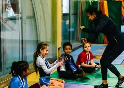 Welfare Officer speaking with smiling children on mats during Chelsea Gymnastics Academy Christmas celebration in Kensington, London.