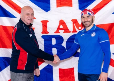 Two smiling coaches shaking hands in front of a British Gymnastics flag during Chelsea Gymnastics Academy Christmas event.
