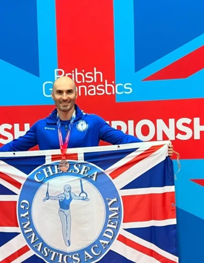 Coach Stefan Kolimechkov holding the Kensington & Chelsea Gymnastics Academy flag and his bronze medal at the 2023 British Gymnastics Adult Championships.