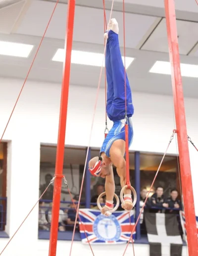 Coach Stefan Kolimechkov holding a controlled handstand on rings during the British Gymnastics Championships 2023, with KCGA flag in the background.
