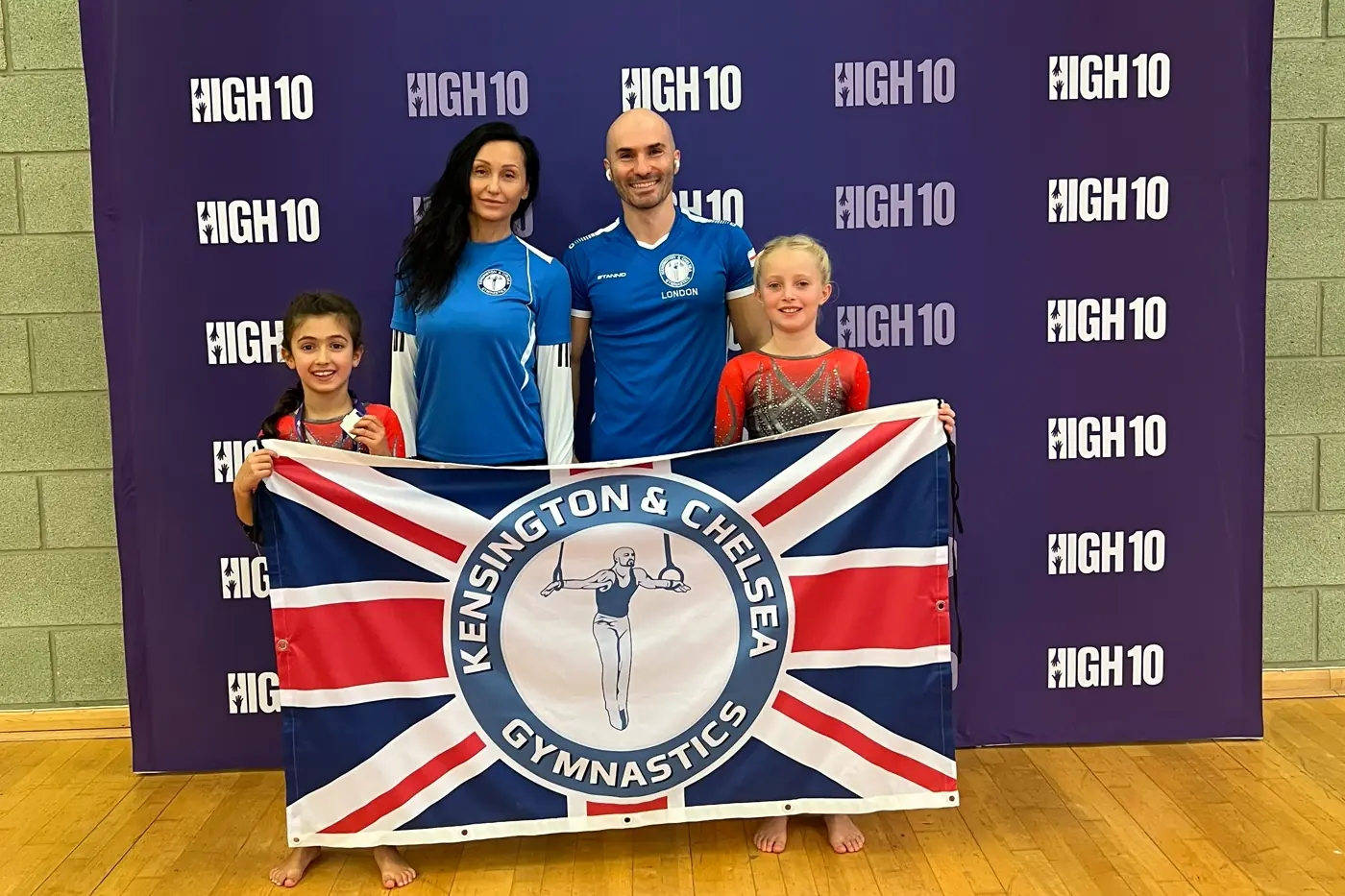 KCGA coaches and young gymnasts holding the Kensington & Chelsea Gymnastics Academy flag at the High Ten Essex Invitational 2025.