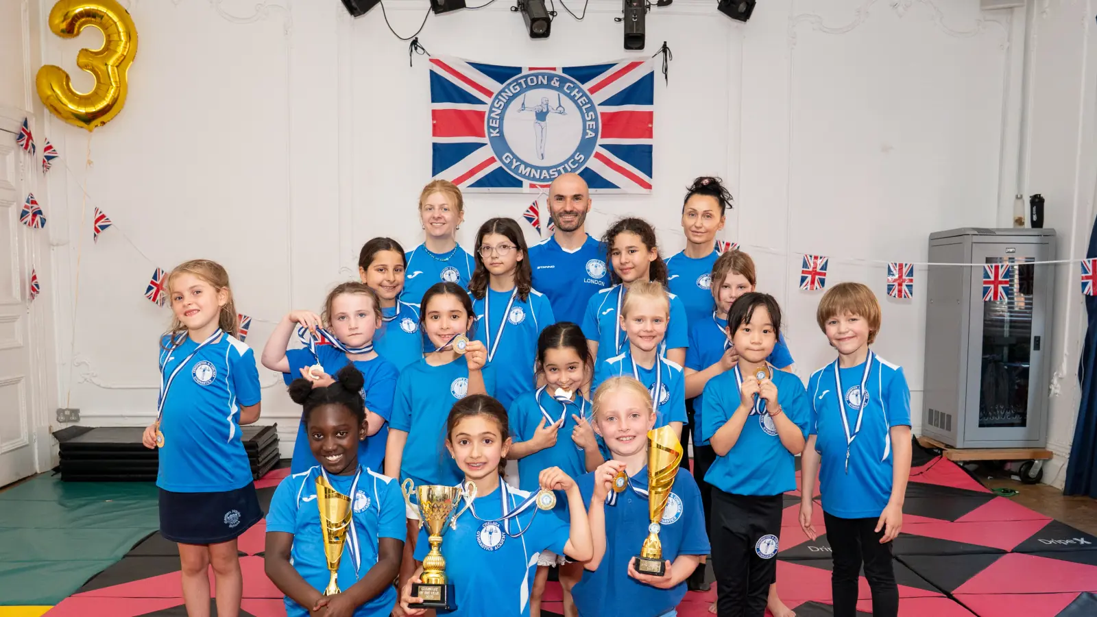 Group photo of children at Chelsea Gymnastics celebrating their achievements with medals and trophies, alongside their coaches from Kensington & Chelsea Gymnastics.