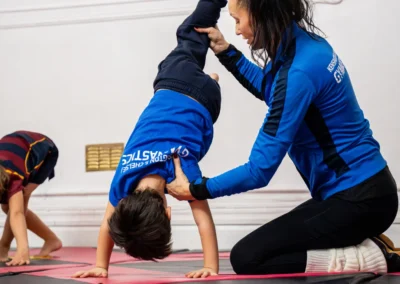 Gymnastics coach providing hands-on support to a child during a controlled skill progression on mats.