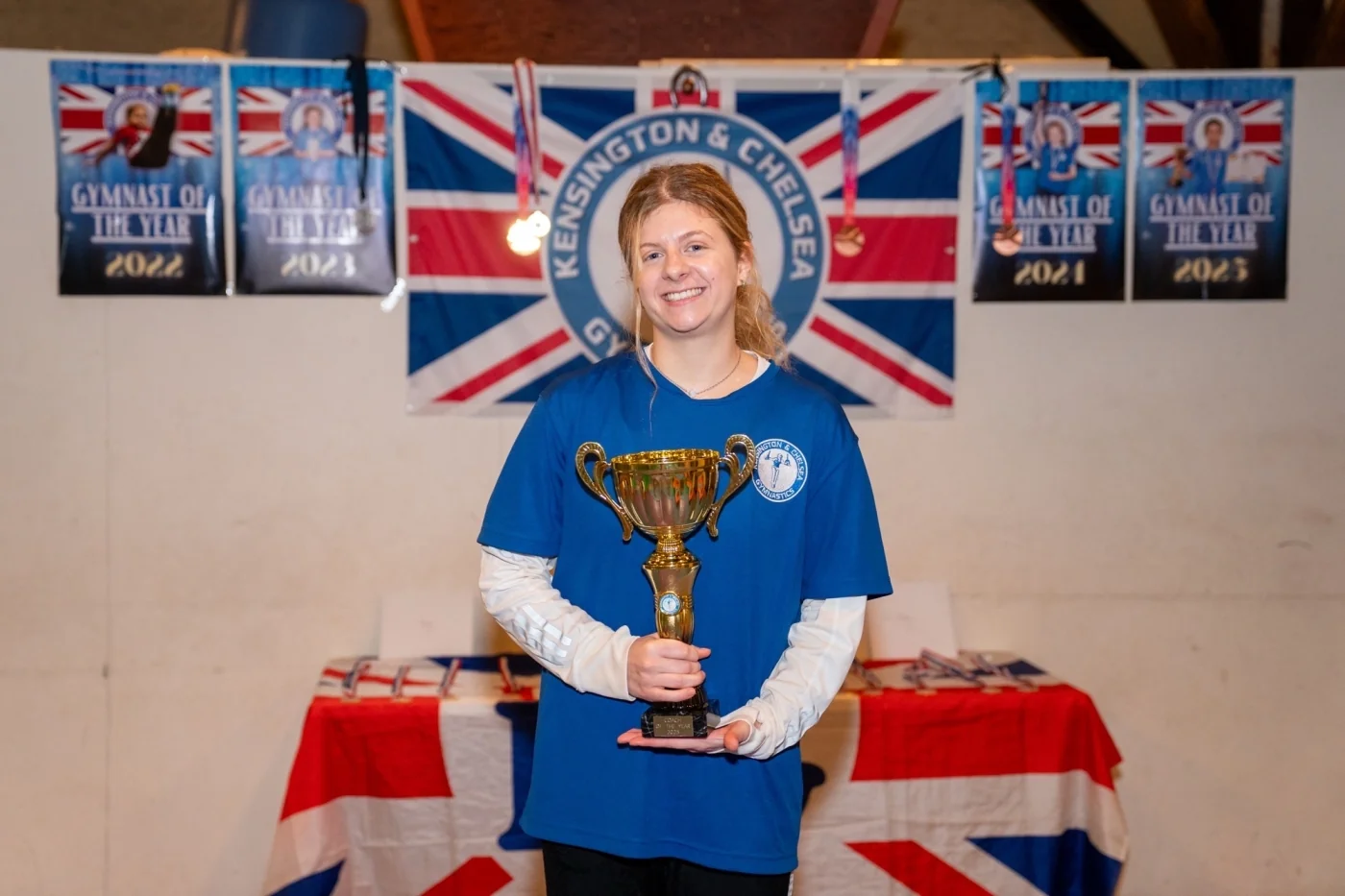 Coach Venia holding the Coach of the Year 2025 trophy at Kensington & Chelsea Gymnastics Academy in London.