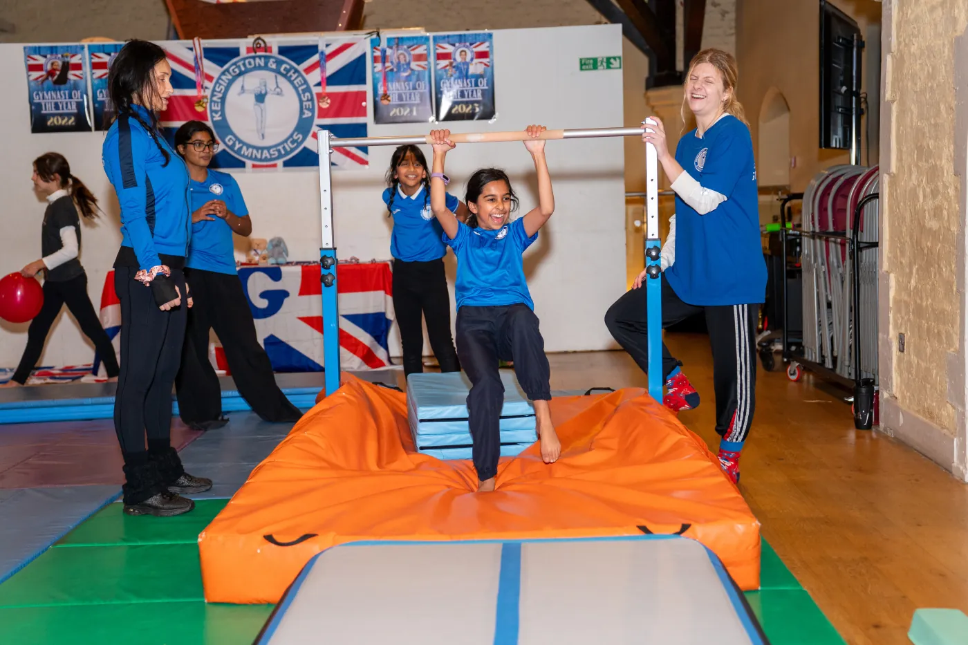 Children practising gymnastics on bars with safety mats and close coach supervision in a recreational class.