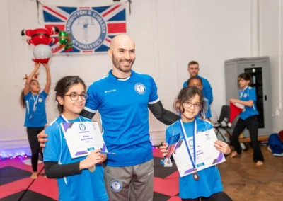Coach with two Knightsbridge gymnasts holding certificates and trophy at KCGA Christmas 2024