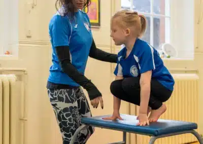 Young girl practising a squat on on gymnastics vault with coach support during Easter 2026 in Chelsea, London SW3