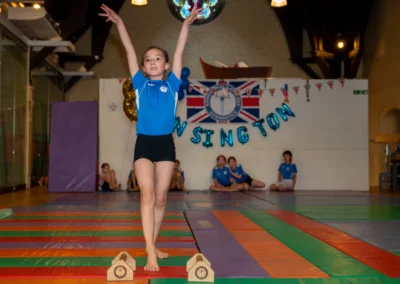 Young girl gymnast raising her arms while balancing beside hand blocks during the KCGA Annual Performance 2025 in Kensington, London.