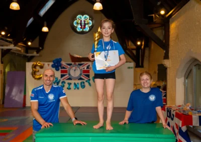 Young gymnast standing with her trophy, medal and certificate alongside KCGA coaches at the Annual Performance 2025 in Kensington, London.