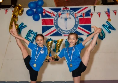 Two young gymnasts holding trophies while performing flexibility poses at the KCGA Annual Performance 2025 in Kensington, London.