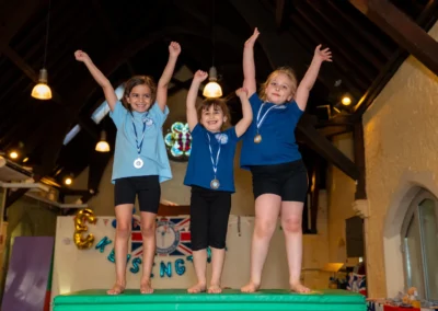 Three young gymnasts wearing medals and standing on a mat with their arms raised at the KCGA Annual Performance 2025 in Kensington, London.