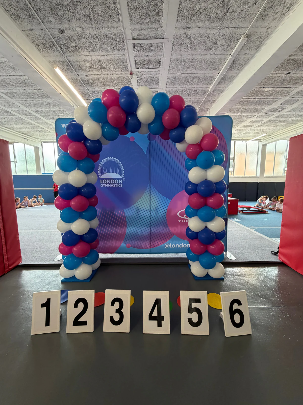 London Gymnastics presentation backdrop, balloon arch and six category markers at the July 2026 pilot.