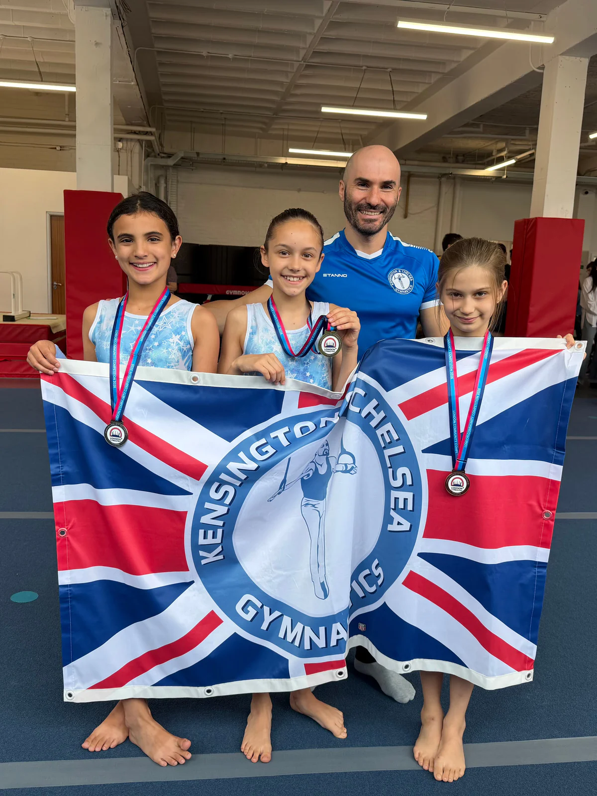 Three young KCGA gymnasts wearing medals with their coach and club flag after the London Community Competition.