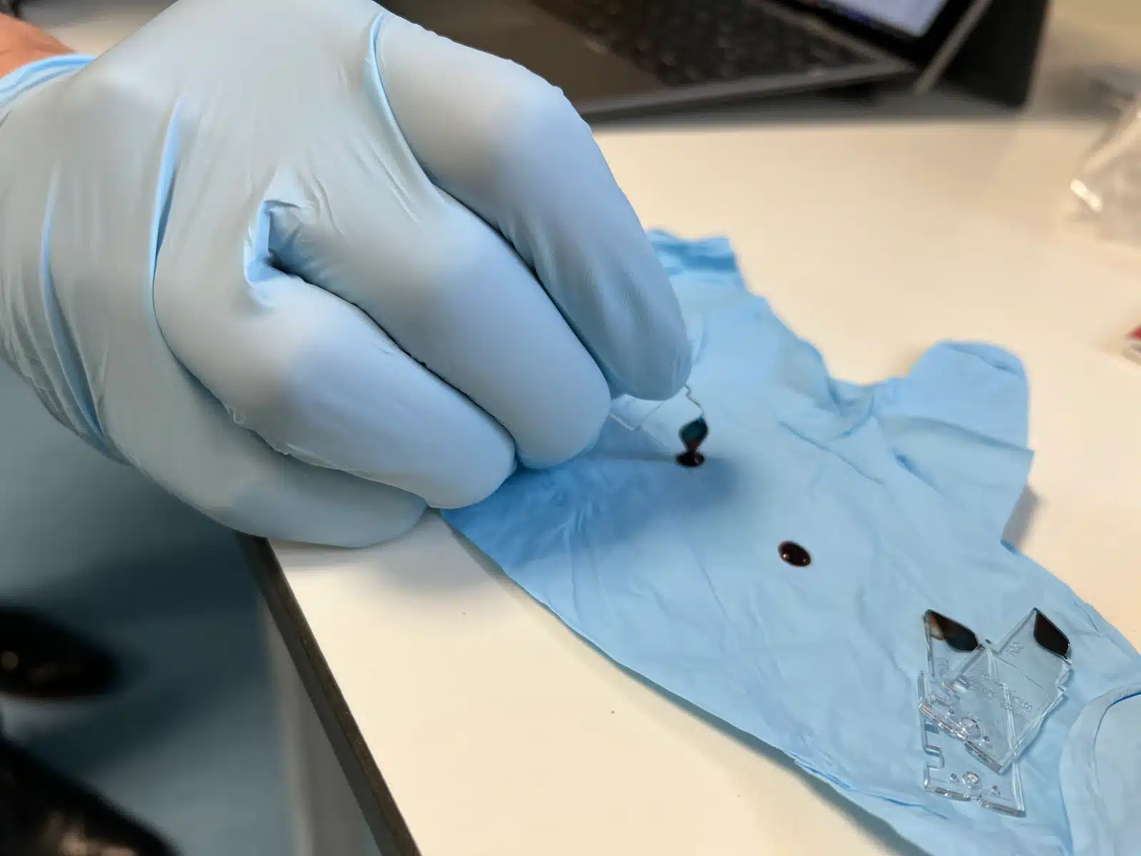 Close-up of a gloved hand carrying out a small blood-sample measurement procedure on a laboratory surface.