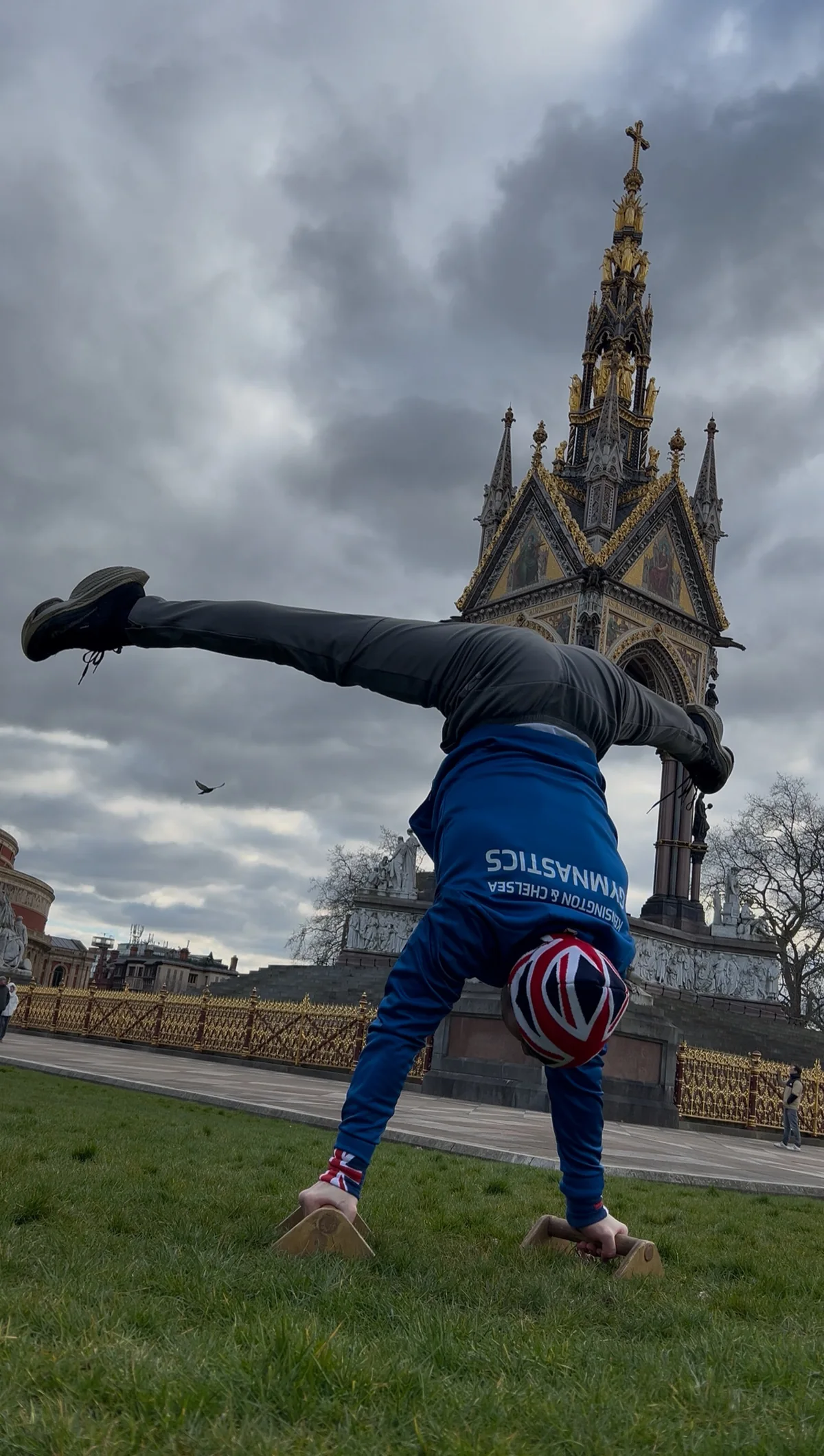 Stefan Kolimechkov performing a press to handstand on mini bars in Kensington Gardens with the Albert Memorial behind.